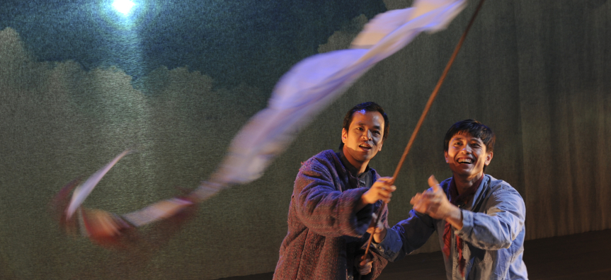 Two men stand side by side - the first is waving a long red flag on a brown stick, the second smiles gleefully looking and pointing at the flag. They stand against a backdrop of a full moon rising over clouds. 